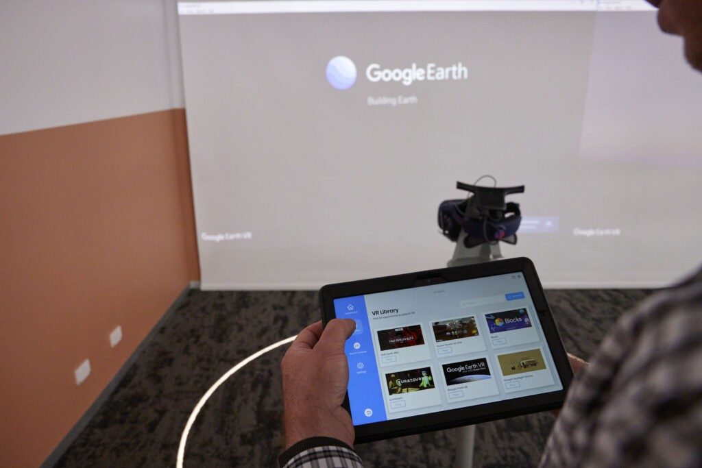Over the shoulder POV of someone holding a tablet, with a projected screen in the background